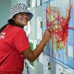 A person wearing a red shirt and a black-and-white checkered hat smiles at the camera while pointing at a map covered in red strings, which are connected to various points. The map and several notes are displayed on a gray bulletin board.