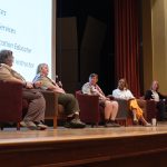 Five individuals are seated on stage in a panel discussion setup. Each person has a microphone. The background includes a large screen displaying text. The panelists appear engaged in conversation, with the audience out of view. The event takes place in an auditorium.